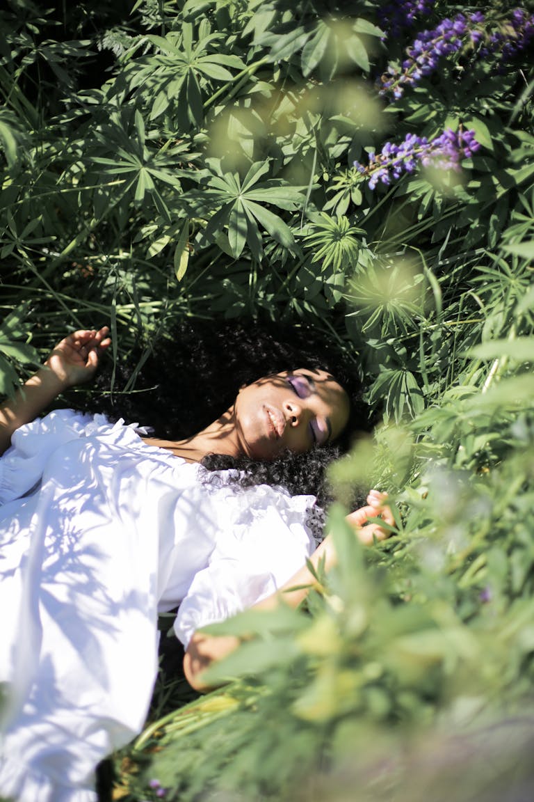 A peaceful woman in a white dress lying among lush greenery and flowers, eyes closed in serenity.