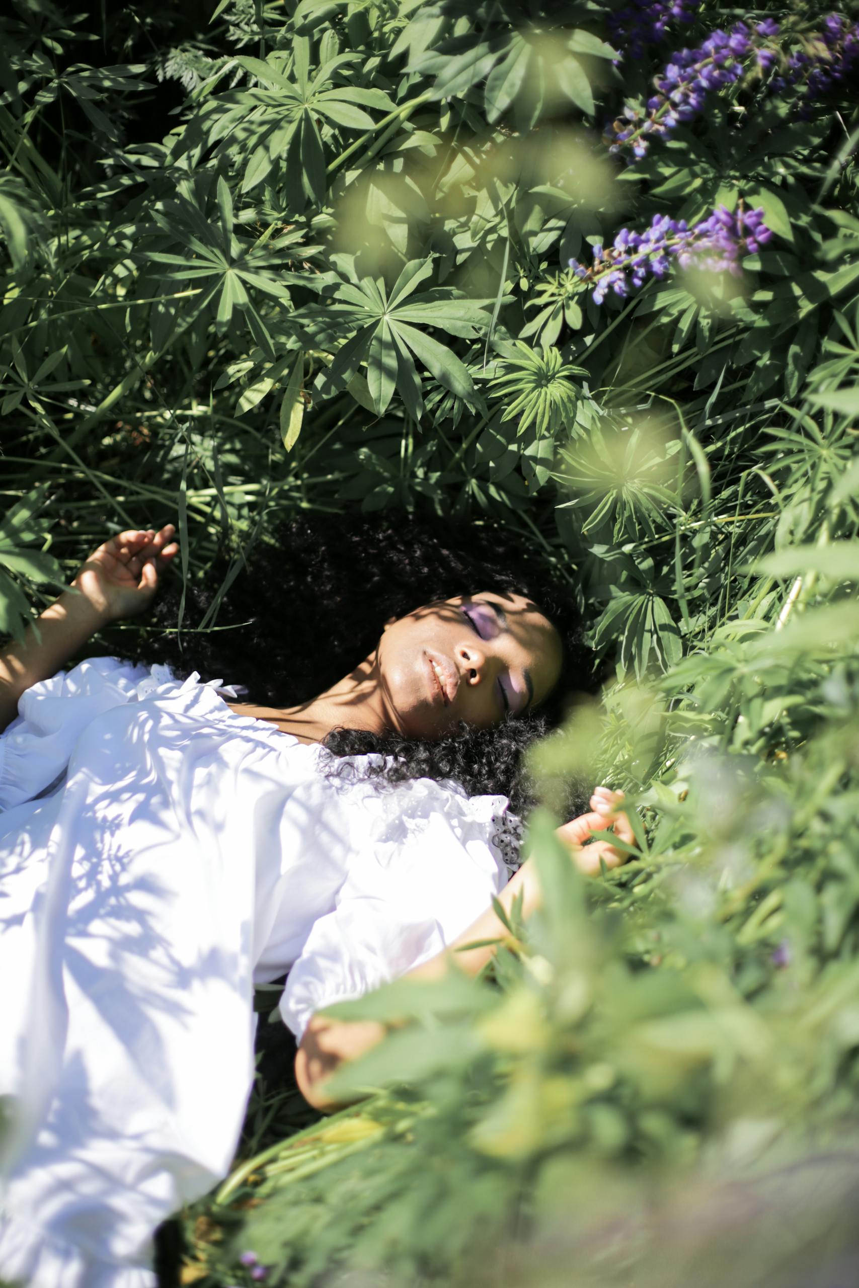 A peaceful woman in a white dress lying among lush greenery and flowers, eyes closed in serenity.
