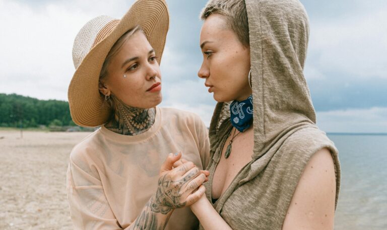 Two women with tattoos, sharing a heartfelt moment on a beach, portraying love and friendship.