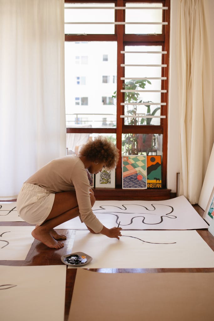 A female artist draws on large white paper in a bright studio, surrounded by wooden floors and paintings.