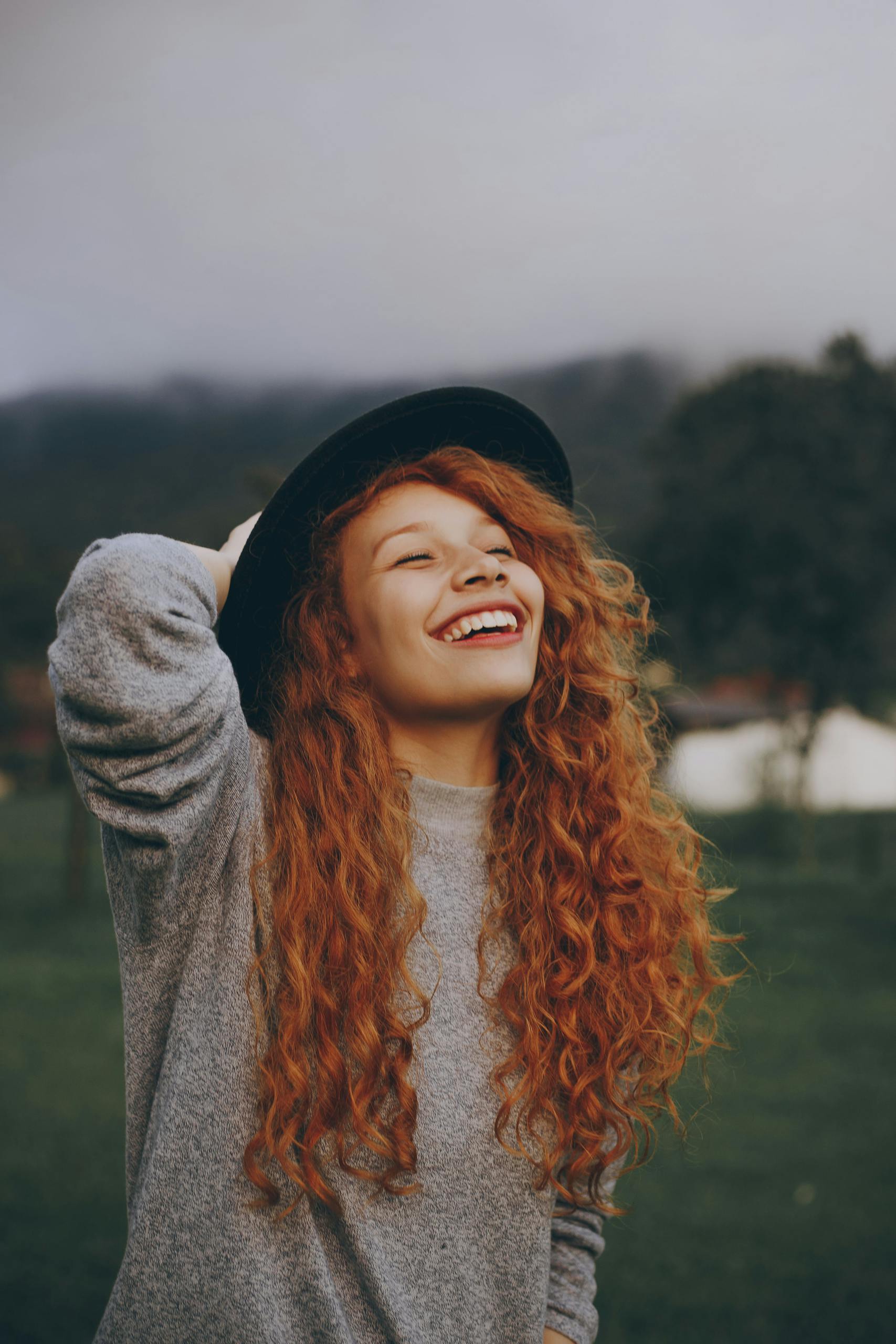 A happy young woman with curly red hair and a hat smiling in a lush green park.