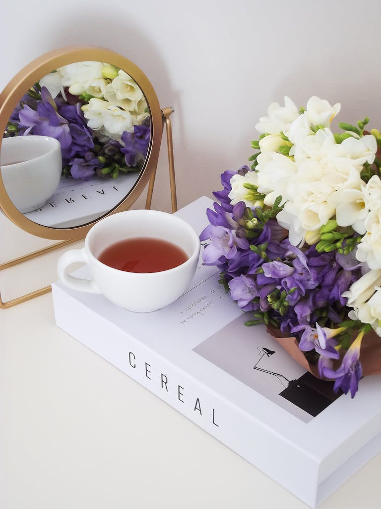 A serene arrangement of a tea cup with violet and white flowers reflected in a mirror, on a book.