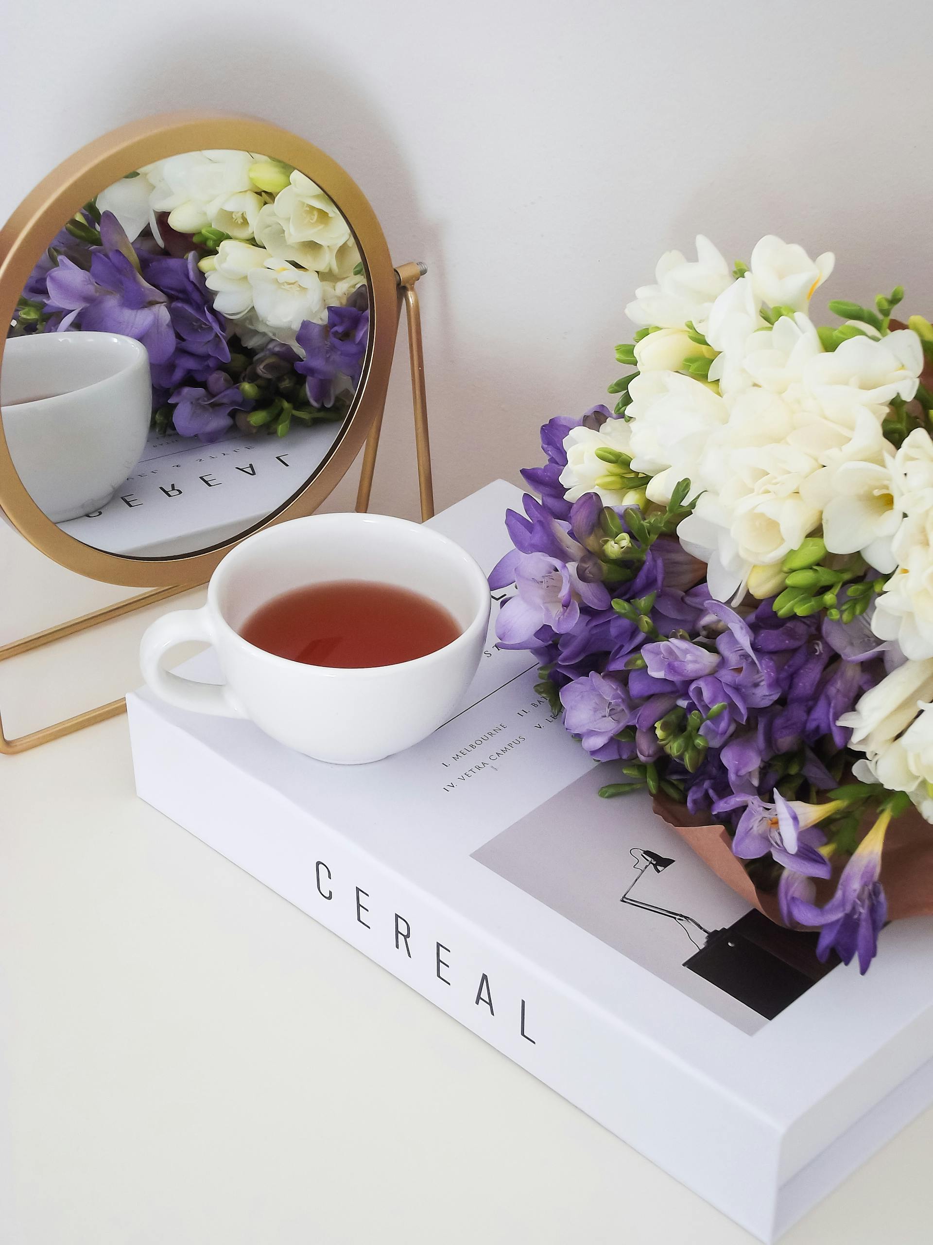 A serene arrangement of a tea cup with violet and white flowers reflected in a mirror, on a book.