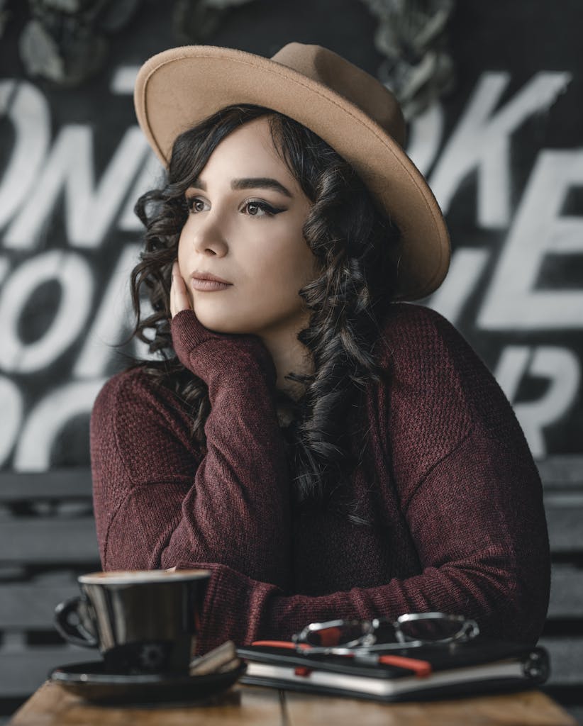 Stylish woman in a brown hat sitting at a cafe table, thoughtful and relaxed.