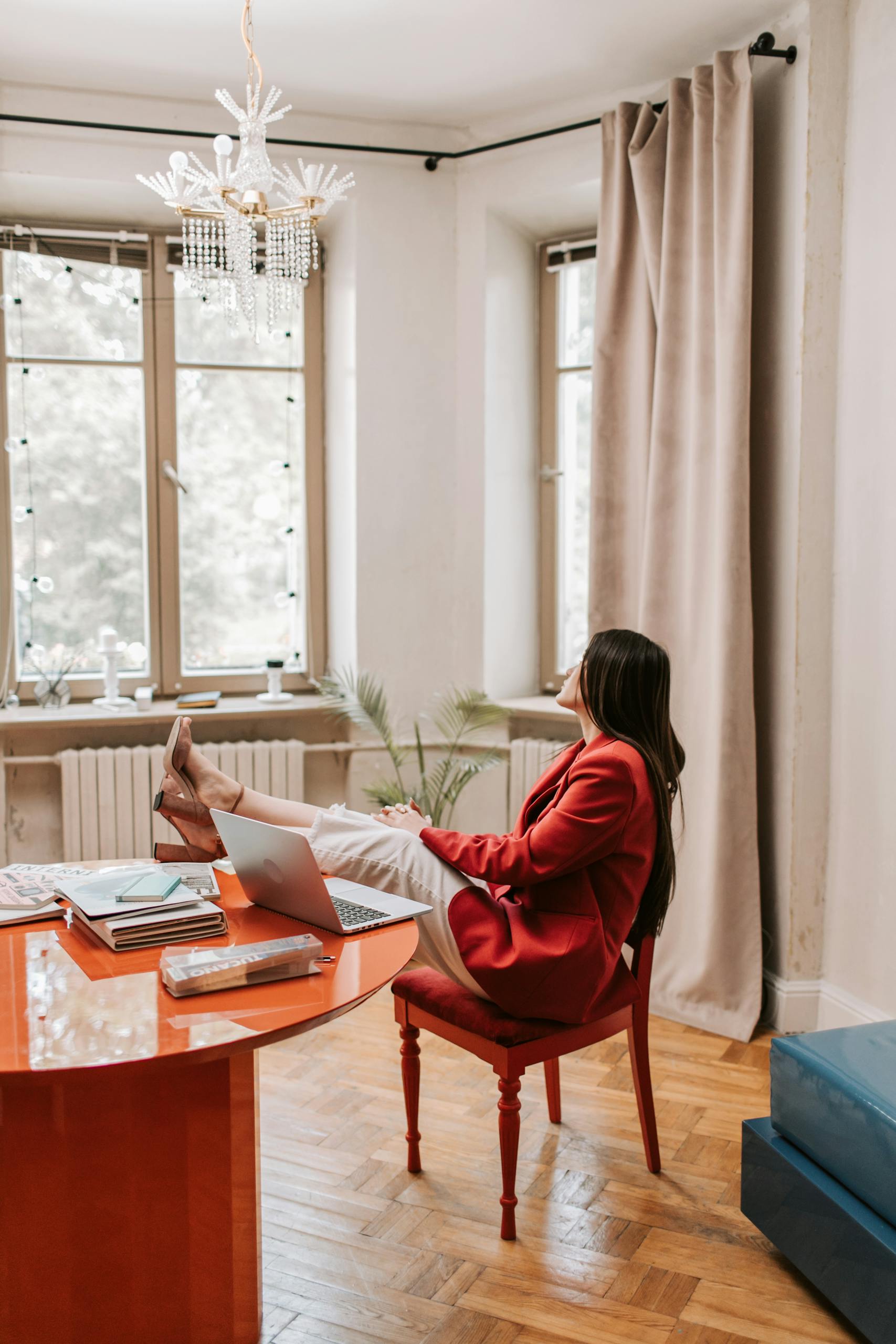 Woman in red blazer relaxing at a desk with a laptop, enjoying a break in a modern office.