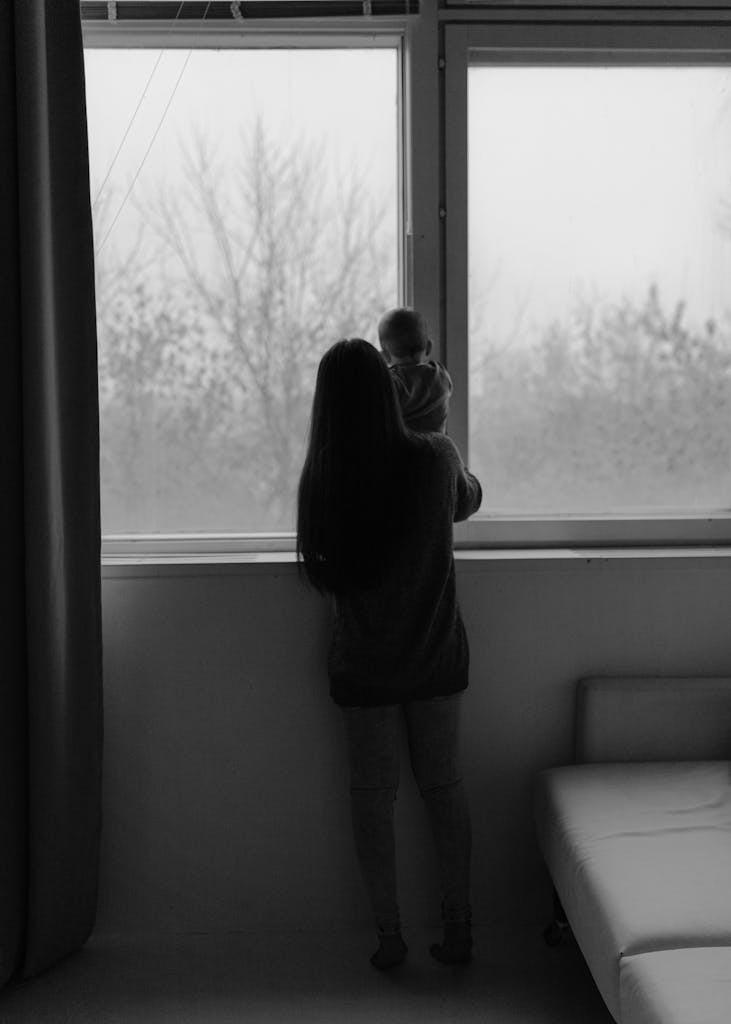 A mother holds her baby while looking out a window, captured in black and white.
