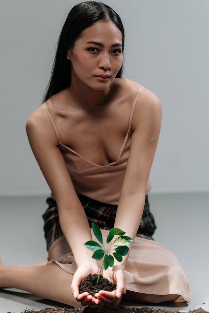 A woman in a spaghetti strap dress holds a small plant in soil, symbolizing eco-consciousness.