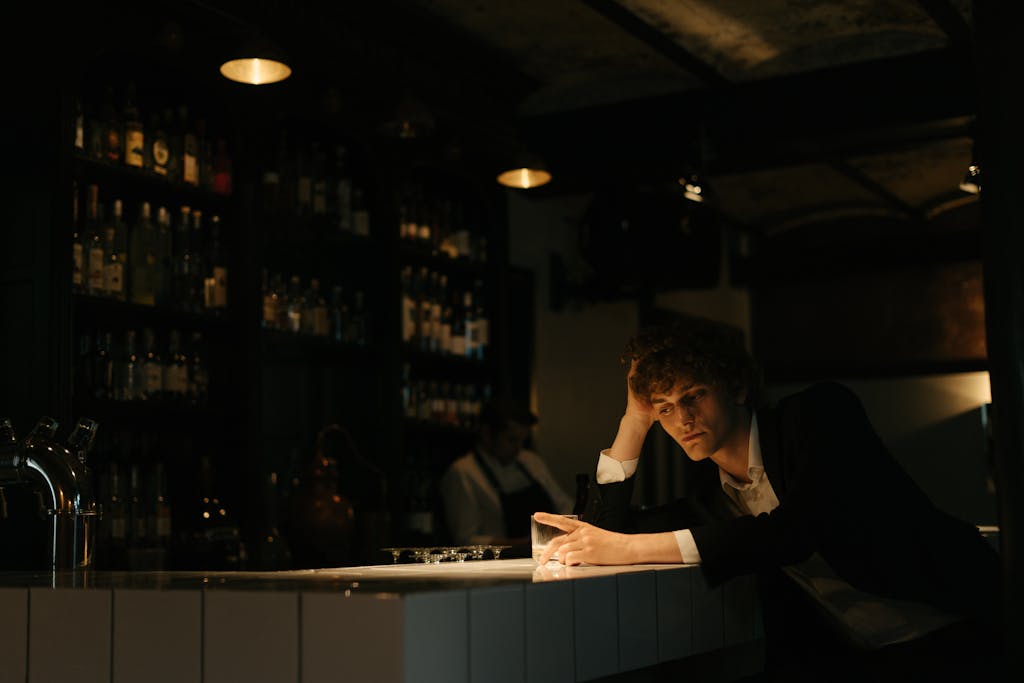 A man wearing a black suit sits alone at a dimly lit bar, looking thoughtful and elegant.